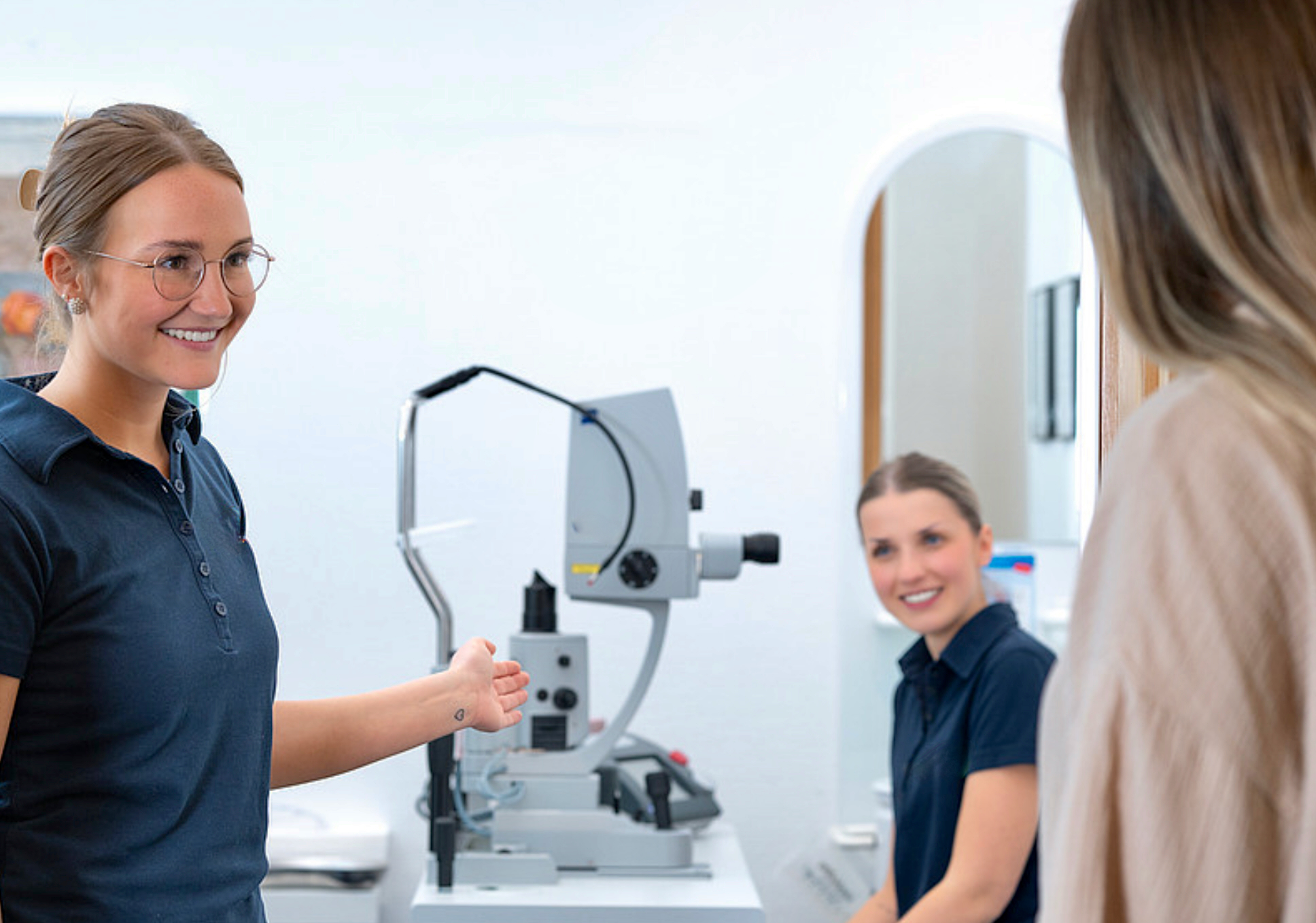 Junge medizinische Mitarbeiterin zeigt Patientin ein ophthalmologisches Gerät in moderner Arztpraxis - Ärztestellen mit Technikbezug