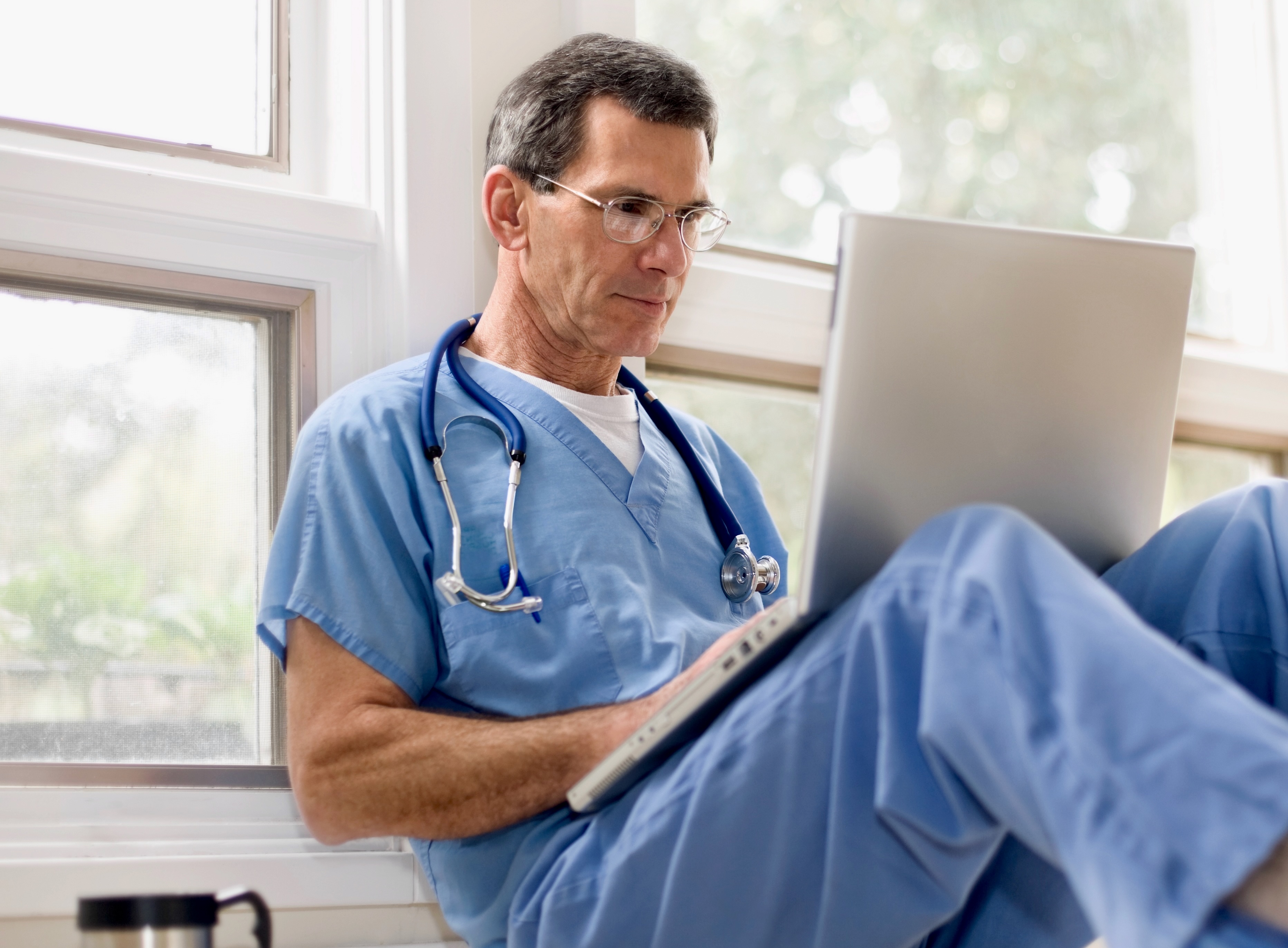 Erfahrener Arzt in blauer Kleidung arbeitet konzentriert am Laptop - Symbol für digitale Arztpraxis, medizinische Software und den Einsatz von Künstlicher Intelligenz im Praxisalltag